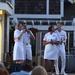 Four individuals dressed in white military-style uniforms are performing on an outdoor stage, with two of them holding microphones and appearing to sing. The performance is taking place at what appears to be an outdoor venue with a shingled building in the background and a band setup visible behind them. An audience is visible in the foreground watching the performance.