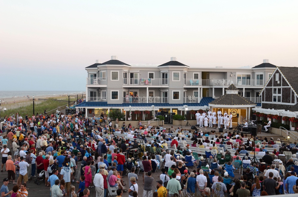 A large outdoor public gathering is taking place on a boardwalk in what appears to be a coastal beach town at dusk. A performance or ceremony is occurring on a stage/gazebo area in front of a multi-story beachside hotel or resort, with performers dressed in white uniforms visible on stage. Hundreds of spectators are gathered, some seated in folding chairs and others standing, watching the event.