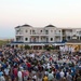 A large outdoor public gathering is taking place on a boardwalk in what appears to be a coastal beach town at dusk. A performance or ceremony is occurring on a stage/gazebo area in front of a multi-story beachside hotel or resort, with performers dressed in white uniforms visible on stage. Hundreds of spectators are gathered, some seated in folding chairs and others standing, watching the event.