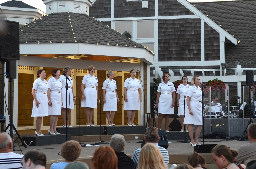 A group of women dressed in white military-style uniforms are performing as a vocal ensemble on an outdoor stage, likely a U.S. Navy choir based on their uniforms. The performance is taking place at what appears to be a gazebo-style outdoor venue decorated with string lights, with a live drummer visible in the background. An audience is seated in the foreground watching the performance.