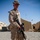 A young male service member in desert camouflage uniform stands at attention on a gravel surface at what appears to be a forward operating base. He is holding a rifle and wearing a boonie hat, with military tents and an Afghan flag visible in the background. The setting and uniform are consistent with a U.S. Marine Corps deployment in Afghanistan.