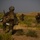Three heavily equipped soldiers are conducting a patrol or tactical movement through an arid, scrub-covered terrain. The soldiers are in a dispersed formation, with at least one appearing to aim a rifle toward a distant objective. The environment features flat, sandy ground with low vegetation and earthen structures visible on the horizon under an overcast sky.