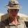 A young male service member wearing USMC MARPAT desert camouflage uniform and a boonie hat is photographed in an arid, forward operating base environment. He appears to be standing outdoors under clear blue skies with military structures visible in the background. His expression is serious and focused.