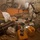 A soldier in full combat gear and helmet is searching through a cluttered storage room or compound interior. The space contains numerous items including containers, sacks, cylinders, and various materials scattered across the floor. A bicycle is visible leaning against the wall in the background.