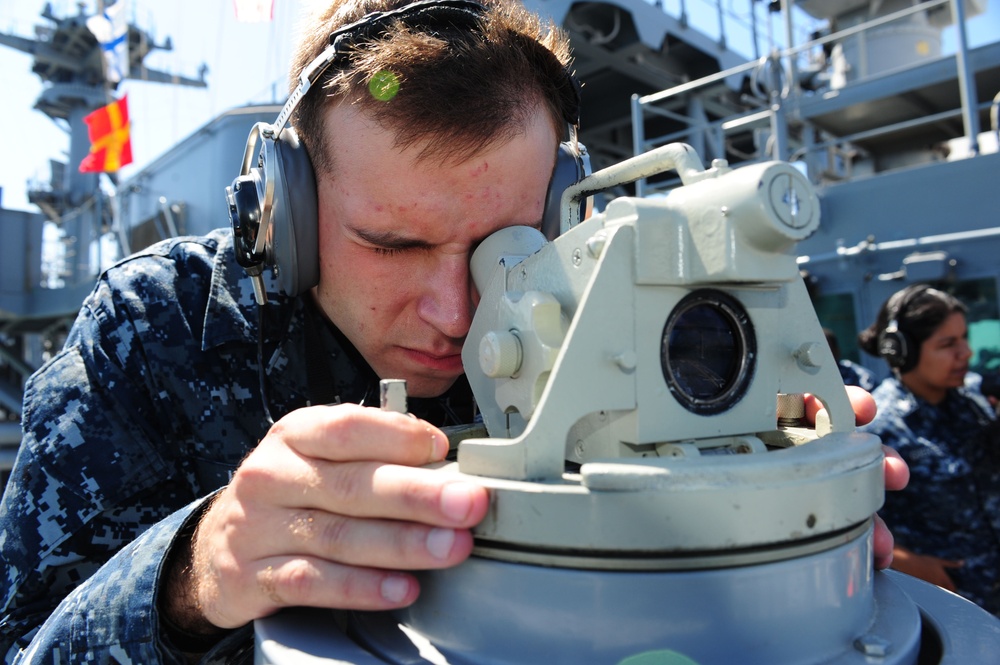 A U.S. Navy sailor in digital camouflage (NWU Type I) uniform is looking through a bearing circle or alidade navigation instrument aboard a naval vessel. The sailor is wearing sound-powered telephone headphones, indicating active communication during a navigation evolution. A second sailor in the same uniform is visible in the background, also wearing headphones.