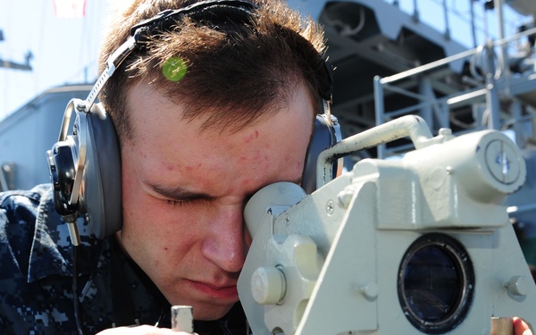 A U.S. Navy sailor in digital camouflage (NWU Type I) uniform is looking through a bearing circle or alidade navigation instrument aboard a naval vessel. The sailor is wearing sound-powered telephone headphones, indicating active communication during a navigation evolution. A second sailor in the same uniform is visible in the background, also wearing headphones.