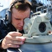 A U.S. Navy sailor in digital camouflage (NWU Type I) uniform is looking through a bearing circle or alidade navigation instrument aboard a naval vessel. The sailor is wearing sound-powered telephone headphones, indicating active communication during a navigation evolution. A second sailor in the same uniform is visible in the background, also wearing headphones.