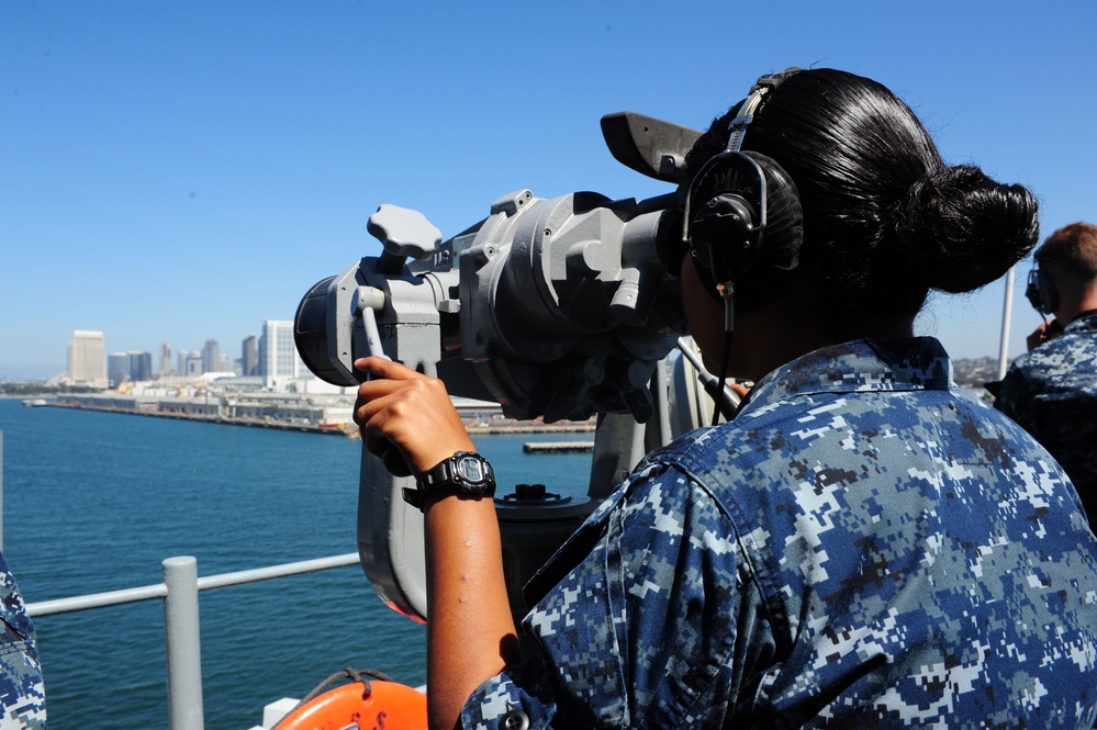 A U.S. Navy sailor in Navy Working Uniform (NWU) Type I digital camouflage is using large mounted binoculars (Big Eyes) aboard a naval vessel while wearing a communications headset. The scene appears to be in a harbor, with a city skyline visible in the background across the water, consistent with San Diego, California. A second sailor in the same uniform is partially visible in the background.