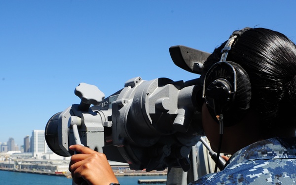 A U.S. Navy sailor in Navy Working Uniform (NWU) Type I digital camouflage is using large mounted binoculars (Big Eyes) aboard a naval vessel while wearing a communications headset. The scene appears to be in a harbor, with a city skyline visible in the background across the water, consistent with San Diego, California. A second sailor in the same uniform is partially visible in the background.