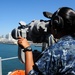 A U.S. Navy sailor in Navy Working Uniform (NWU) Type I digital camouflage is using large mounted binoculars (Big Eyes) aboard a naval vessel while wearing a communications headset. The scene appears to be in a harbor, with a city skyline visible in the background across the water, consistent with San Diego, California. A second sailor in the same uniform is partially visible in the background.
