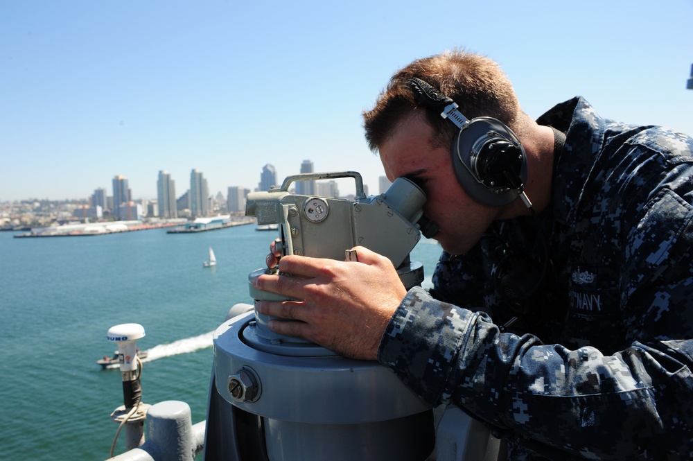 A U.S. Navy sailor in digital camouflage (NWU Type I) uniform is using a bearing circle or alidade optical instrument on the bridge wing of a naval vessel. The sailor is wearing sound-powered telephone headsets, indicating active communication during navigation or watch duties. The background reveals a harbor cityscape consistent with San Diego, California, with the downtown skyline visible across the water.