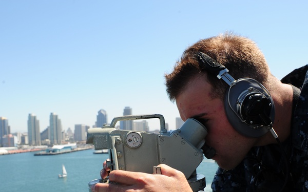 A U.S. Navy sailor in digital camouflage (NWU Type I) uniform is using a bearing circle or alidade optical instrument on the bridge wing of a naval vessel. The sailor is wearing sound-powered telephone headsets, indicating active communication during navigation or watch duties. The background reveals a harbor cityscape consistent with San Diego, California, with the downtown skyline visible across the water.