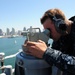A U.S. Navy sailor in digital camouflage (NWU Type I) uniform is using a bearing circle or alidade optical instrument on the bridge wing of a naval vessel. The sailor is wearing sound-powered telephone headsets, indicating active communication during navigation or watch duties. The background reveals a harbor cityscape consistent with San Diego, California, with the downtown skyline visible across the water.