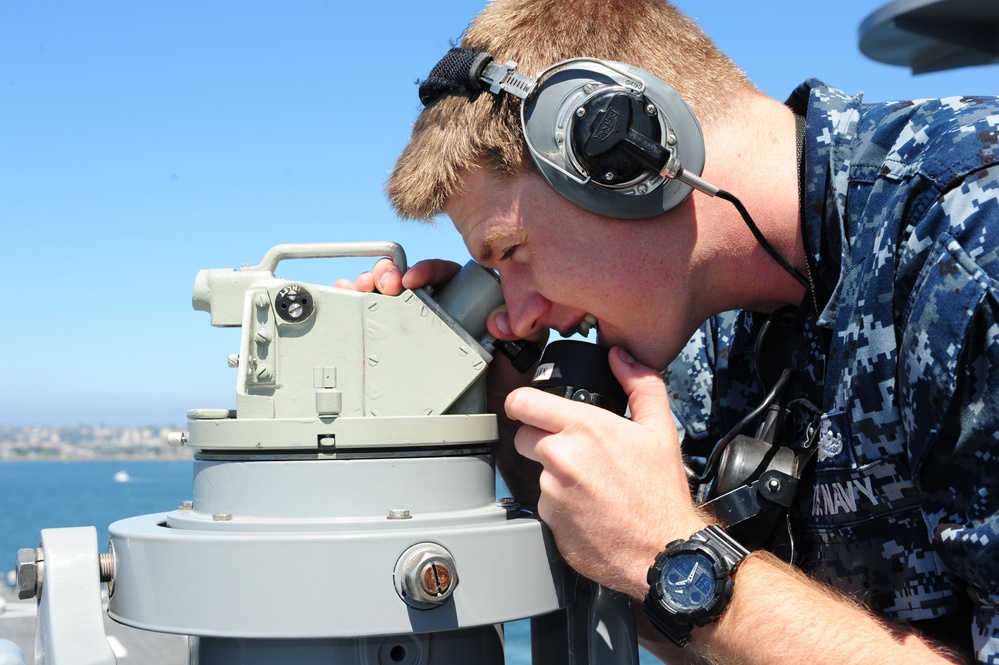 A U.S. Navy sailor in digital camouflage (NWU Type I) uniform is looking through a bearing circle or alidade mounted on a gyrocompass repeater aboard a naval vessel. The sailor is wearing communication headphones and a tactical wristwatch, suggesting active bridge watch duties. The background shows open water and a distant coastline under clear blue skies.