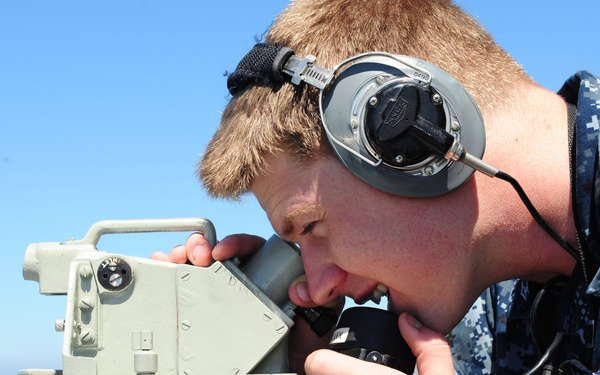 A U.S. Navy sailor in digital camouflage (NWU Type I) uniform is looking through a bearing circle or alidade mounted on a gyrocompass repeater aboard a naval vessel. The sailor is wearing communication headphones and a tactical wristwatch, suggesting active bridge watch duties. The background shows open water and a distant coastline under clear blue skies.