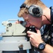 A U.S. Navy sailor in digital camouflage (NWU Type I) uniform is looking through a bearing circle or alidade mounted on a gyrocompass repeater aboard a naval vessel. The sailor is wearing communication headphones and a tactical wristwatch, suggesting active bridge watch duties. The background shows open water and a distant coastline under clear blue skies.