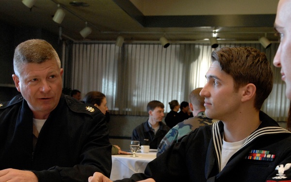 A group of U.S. Navy personnel are seated at a table in what appears to be a conference or meeting room, engaged in conversation. A senior enlisted sailor in a dark service uniform speaks with two junior sailors, one wearing a dress blue uniform with service ribbons and a Seaman rating badge. The setting suggests a mentorship session, briefing, or professional development event.