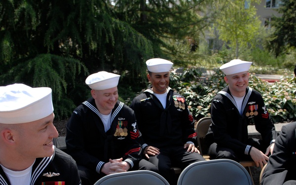 A group of U.S. Navy personnel in dress uniforms are gathered outdoors in what appears to be a courtyard or garden setting. The sailors are seated on folding chairs in a relaxed, informal arrangement, engaged in conversation. The group includes enlisted sailors in crackerjack uniforms with white dixie cup hats and a senior officer in a dark service dress uniform with gold sleeve stripes.