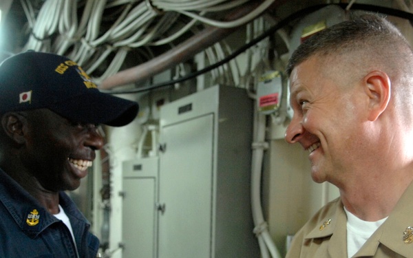Two U.S. Navy personnel are engaged in a friendly conversation aboard what appears to be a naval vessel, both smiling broadly. The sailor on the left wears a dark navy working uniform with a cap bearing a Japanese flag patch and a name tag reading 'DMUSO', while the officer on the right wears a tan service uniform with ribbons, a warfare device, and rank insignia. The setting appears to be an interior compartment of a ship with visible wiring, conduits, and electrical panels in the background.