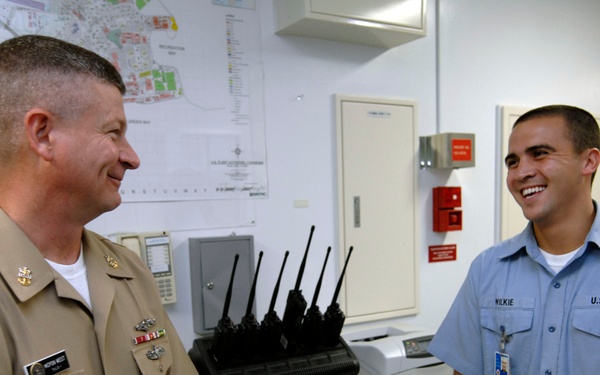 Two U.S. Navy personnel are engaged in a friendly conversation inside what appears to be a military administrative or security office. The individual on the left wears senior enlisted or officer khaki service uniform with ribbons and insignia, while the person on the right wears a blue utility uniform with the name tag 'WILKIE' and a Petty Officer Third Class rank insignia. Both individuals appear relaxed and smiling.