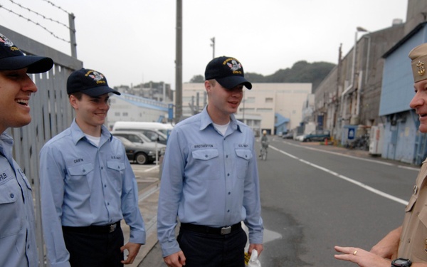 A senior U.S. Navy officer in tan service dress uniform is conversing with three junior enlisted sailors wearing blue utility uniforms and navy ball caps. The interaction appears informal and friendly, taking place outdoors near a gated facility on what appears to be a naval base. The setting, architectural style, and signage suggest this may be a U.S. naval installation in Japan.