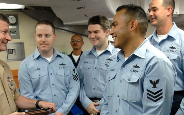 A U.S. Navy officer in khaki service uniform is presenting an award or plaque to enlisted sailors wearing blue utility uniforms. The ceremony appears to be taking place aboard a naval vessel in an interior compartment. The sailors are smiling and appear to be in good spirits during what seems to be a recognition or award ceremony.