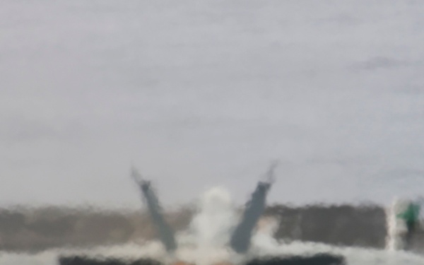A twin-engine carrier-based fighter jet, consistent with an F/A-18 Hornet or Super Hornet, is photographed head-on on what appears to be an aircraft carrier flight deck. The aircraft's engines are visibly lit with afterburner glow, suggesting it is in a pre-launch or run-up phase. Steam or jet wash is visible around the aircraft, consistent with catapult launch operations.