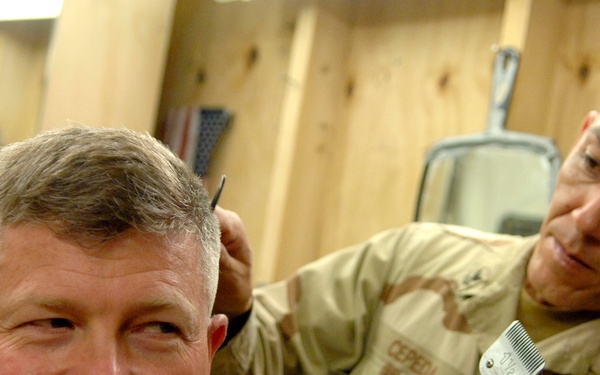 A U.S. Navy servicemember in desert camouflage uniform is giving a haircut to another individual seated in a barber chair. The barber is wearing a name tape reading 'CEPEDA' and a 'NAVY' branch tape, and is using electric clippers. The setting appears to be a makeshift or forward-deployed barbershop constructed with plywood walls.