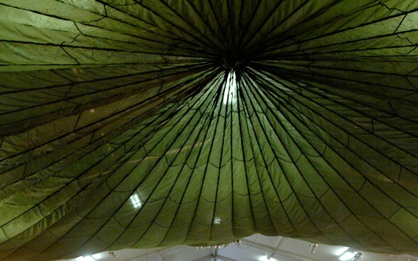 A military briefing or presentation is taking place inside a large tent structure, likely at a forward operating base. A single uniformed individual stands at the front before a projection screen, addressing a seated audience of military personnel in desert camouflage uniforms. The tent features a distinctive green camouflage netting ceiling draped over a white inner tent structure with fluorescent lighting.