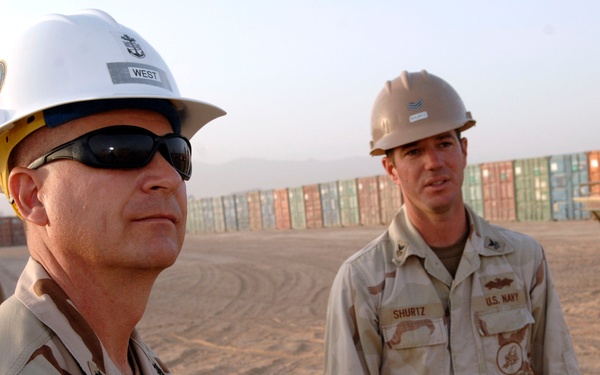 A group of U.S. Navy Seabee personnel are gathered in a sandy, arid environment, wearing desert camouflage uniforms and hard hats. The foreground figure wears a white hard hat labeled 'WEST' and dark sunglasses. Shipping containers are visible stacked in the background, suggesting a forward operating base or port facility.