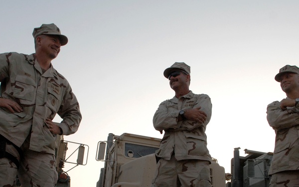 Three military personnel in desert camouflage uniforms (DCU pattern) stand in a relaxed posture in front of military vehicles, likely at a forward operating base or staging area. The setting appears to be a arid/desert environment during early morning or evening based on the low light conditions. The soldiers appear to be engaged in casual conversation.