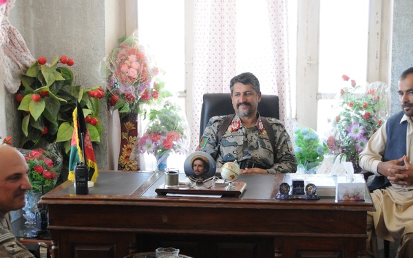 The image shows a formal meeting in what appears to be an official office, with four individuals seated in a discussion setting. Two individuals wearing US military camouflage uniforms are meeting with a person in Afghan military/police uniform seated behind a large wooden desk, and a civilian in traditional Afghan attire. The office is decorated with numerous artificial flower arrangements and an Afghan flag is visible in the background.