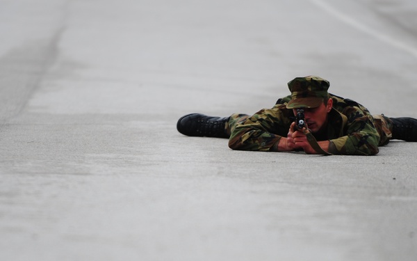 A soldier in camouflage uniform is lying prone on a concrete surface, aiming a firearm toward the camera. The individual is wearing a camouflage cap, matching battle dress uniform (BDU), and black boots. The setting appears to be a paved area such as a runway, road, or training ground.