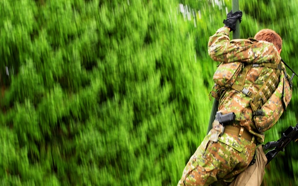 A soldier in camouflage uniform is rappelling or climbing down a rope from an elevated structure, carrying full combat gear including a rifle. The image is captured with motion blur applied to the background, suggesting a panning camera technique that emphasizes movement. The setting appears to be a military training facility surrounded by dense green trees.
