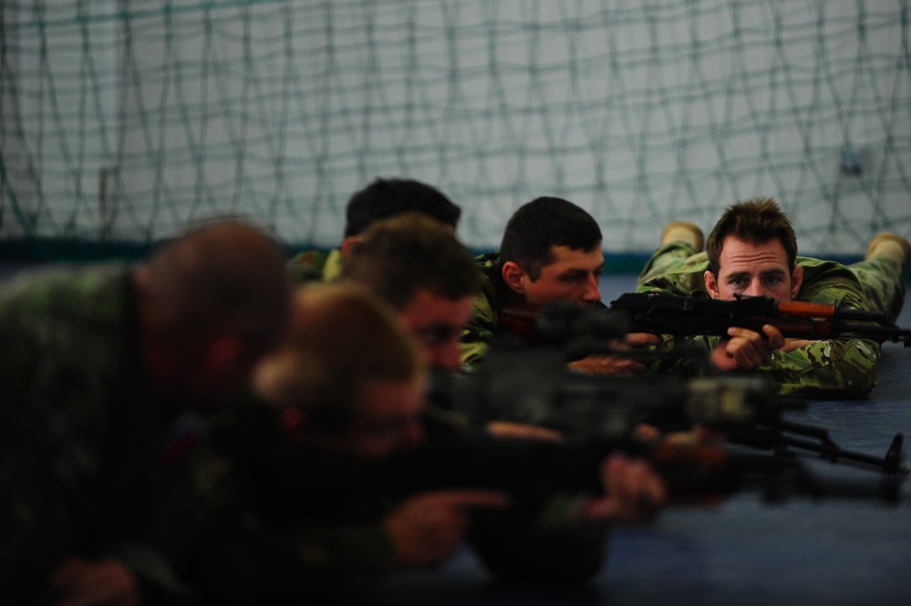 A group of military personnel in camouflage uniforms are lying prone in a line, aiming rifles in a training exercise conducted indoors. The setting appears to be an indoor shooting range or training facility, identifiable by the netting visible in the background. The image uses shallow depth of field, with the foreground figures blurred and the focus on the middle-to-right subjects.