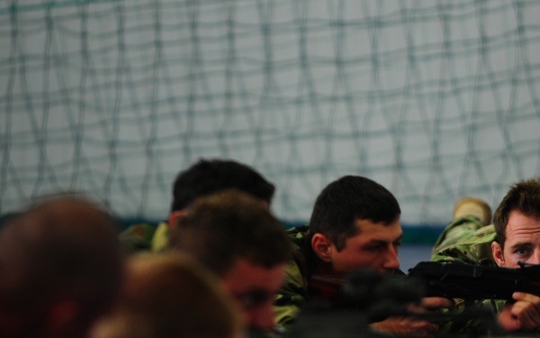 A group of military personnel in camouflage uniforms are lying prone in a line, aiming rifles in a training exercise conducted indoors. The setting appears to be an indoor shooting range or training facility, identifiable by the netting visible in the background. The image uses shallow depth of field, with the foreground figures blurred and the focus on the middle-to-right subjects.