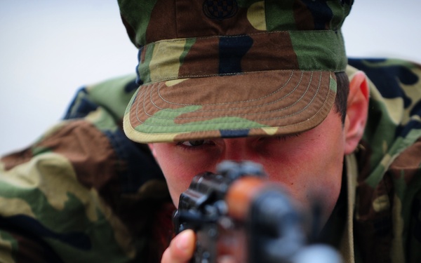 A soldier in woodland camouflage uniform is in a prone shooting position, aiming a firearm directly toward the camera. The image is taken from a low angle, placing the viewer along the barrel's line of sight. The close-up composition emphasizes the shooter's focused expression and weapon handling technique.