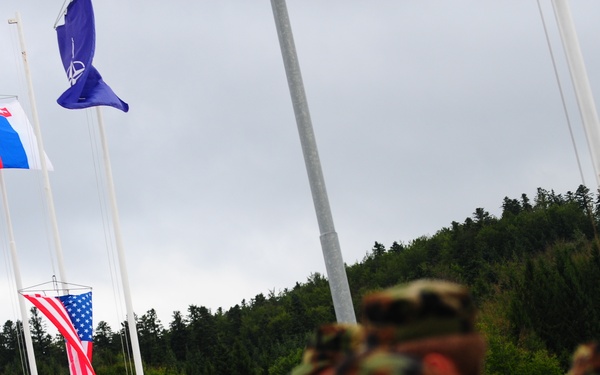 A group of military personnel in camouflage uniforms are assembled in formation, likely during a multinational military ceremony or exercise. Multiple national and organizational flags are visible on flagpoles in the background, including the NATO flag, the United States flag, the Slovak flag, and what appears to be the Estonian flag. The setting appears to be an outdoor military installation surrounded by forested hills.