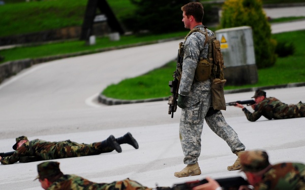 A military training exercise is taking place on a paved surface, likely a base or training facility. A standing soldier in US Army ACU (Army Combat Uniform) with tan boots and a tactical backpack appears to be supervising or instructing several soldiers in woodland camouflage uniforms who are in prone firing positions. The prone soldiers are aiming rifles in a coordinated direction, suggesting a drill or training scenario.