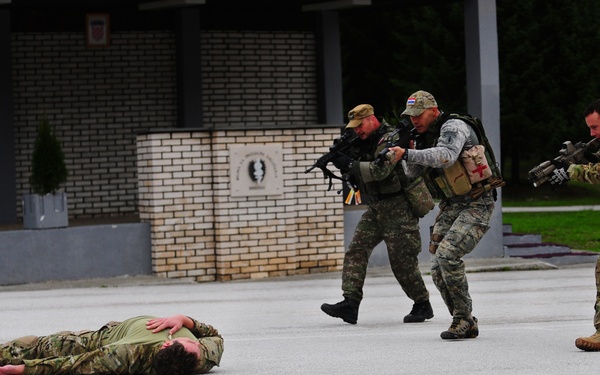 The image depicts what appears to be a military training exercise or demonstration on a paved area near a brick building. Three armed soldiers in tactical gear are advancing toward a fourth soldier lying face-down on the ground. One of the standing soldiers displays a red cross medical patch, suggesting a combat casualty response scenario.