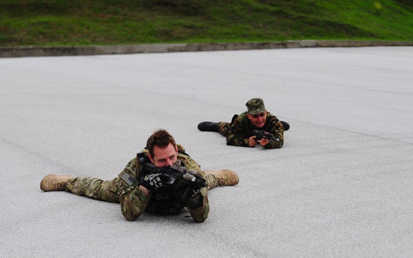 Four military personnel in camouflage uniforms are lying prone on a large paved surface, aiming rifles in a low-crawl tactical position. The foreground soldier wears a multicam/OCP-style uniform with what appears to be a US flag patch, while the others wear woodland camouflage. This appears to be a military training exercise conducted on an open concrete or asphalt area.
