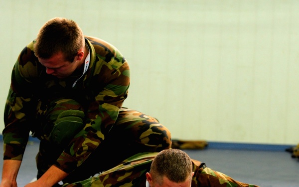 Two individuals wearing matching woodland camouflage uniforms are engaged in what appears to be a military or law enforcement combatives training exercise on a blue mat floor. One person is prone on the ground while the other kneels over them, applying what appears to be a restraint or control technique to the lower body. The setting is an indoor training facility with light-colored walls.