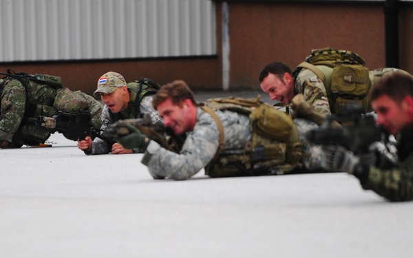 A group of approximately five military personnel in prone firing positions on a hard surface, appearing to conduct a tactical training exercise. The soldiers are wearing various camouflage uniforms and carrying full tactical gear including backpacks and rifles. The setting appears to be an outdoor training area adjacent to a building.