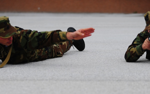 Two soldiers in woodland camouflage uniforms are lying prone on a flat gray surface, practicing tactical drills. The soldier on the left is making a hand signal while holding a rifle with a sling, and the soldier on the right is aiming a pistol forward. The setting appears to be a training exercise on a paved or concrete surface near a brick wall.