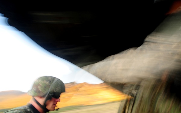 A military personnel wearing camouflage uniform and helmet is visible inside or near a military aircraft, likely during a loading or unloading operation. The image is captured with significant motion blur, suggesting rapid movement or a panning camera technique. The background reveals an open landscape with hills and golden-colored terrain.