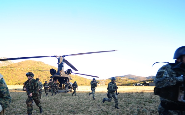 Military personnel are rapidly disembarking from a CH-47 Chinook tandem-rotor helicopter on an open field. Soldiers in camouflage uniforms and full combat gear are running away from the aircraft in what appears to be a tactical insertion exercise or operation. The setting is a rural, hilly landscape under clear blue skies.