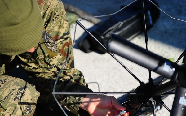 A military personnel wearing digital camouflage uniform and olive green beanie is crouching on a paved surface, assembling or maintaining what appears to be a large multi-rotor drone or UAV (unmanned aerial vehicle). The soldier is working on the central hub of the device, connecting or adjusting wiring and components. Two unit patches are visible on the uniform sleeve.