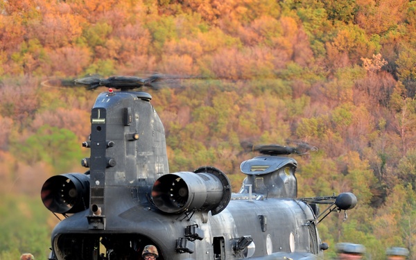 Two dark-colored tandem-rotor helicopters, consistent with CH-47 Chinook variants, are landed in an open field with soldiers rapidly deploying from the rear ramp. Multiple soldiers in camouflage uniforms are exiting and moving away from the aircraft in what appears to be a tactical insertion exercise. The scene takes place against a backdrop of autumn foliage, suggesting a fall training exercise.