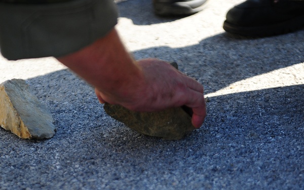 A close-up photograph shows a person's hand placing or picking up a rock on an asphalt surface. Multiple rocks of varying sizes are visible on the pavement, and several people's feet and lower legs are visible in the background. The image is taken at ground level with strong directional sunlight creating shadows.