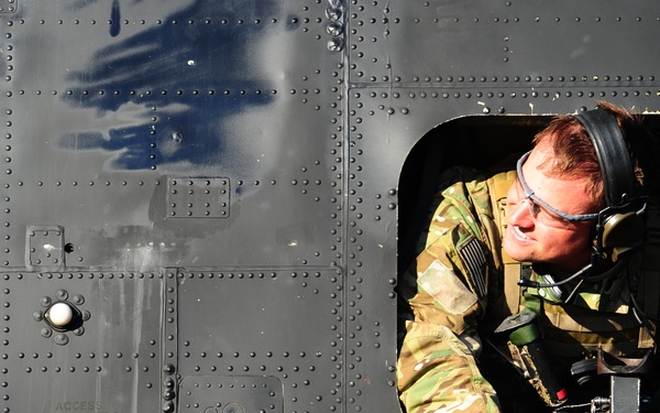 A military personnel member is leaning out of a window or hatch of a military helicopter, wearing camouflage uniform, tactical gloves, protective eyewear, and a communications headset. The aircraft fuselage is dark olive/black with visible riveted metal paneling and stenciled text reading 'ACCESS RAMP FAOL ON STOP UP'. The person appears to be inspecting or monitoring something outside the aircraft.